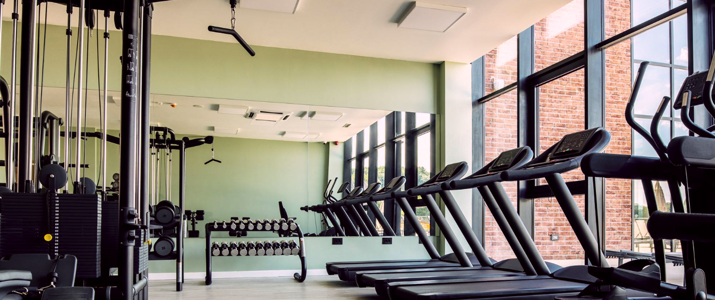 Светлый тренажерный зал с панорамными окнами, возле которых беговые дорожки, кроме них большое количество различных тренажеров, большое зеркало на стене